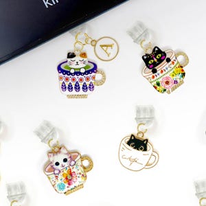 Puede incluir: Una colección de 10 marcapáginas temáticos de gatos coloridos. Cada marcapáginas presenta un diseño de gato diferente, incluyendo gatos en tazas de té, gatos en bombillas y gatos en otros entornos caprichosos. Los marcapáginas están hechos de metal y tienen un acabado dorado. El texto "PAWsitively PAW" es visible en la esquina inferior derecha de la imagen.