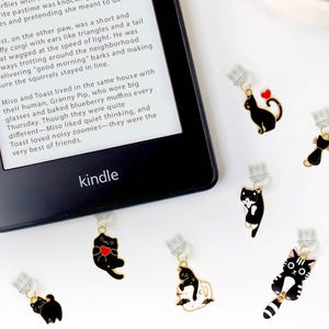 May include: A close-up of a Kindle e-reader with a book about a cat and dog named Miso and Toast.  The image also shows 12 black and white cat bookmarks with gold accents.  The text on the bookmark reads "Paw-lilly Paw".