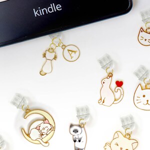 May include: A collection of 15 gold-tone cat-themed bookmark clips. The clips feature various designs, including cats sleeping, drinking coffee, and holding hearts. The clips are all attached to clear plastic holders. The text "PAWilly PAW" is visible in the bottom right corner of the image.