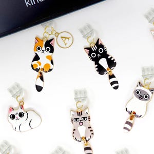 Puede incluir: Un conjunto de 12 clips de marcapáginas con temática de gatos. Los clips están hechos de metal y presentan gatos de diferentes colores con diversas expresiones. El texto "PAWilly PAW" es visible en la esquina inferior derecha de la imagen.