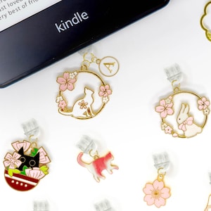 Puede incluir: Un conjunto de diez clips de marcapáginas dorados con varios diseños que presentan gatos, conejos y flores. Los diseños son en rosa, blanco, negro y azul. Los clips están sobre un fondo blanco. El texto "PAWilly PAW" es visible en la esquina inferior derecha.