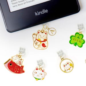Può includere: Una collezione di ciondoli colorati, tra cui gatti fortunati, trifogli e un cappello da folletto. I ciondoli sono attaccati a clip trasparenti, adatti per l'uso come segnalibri o accessori. Un lettore Kindle è sullo sfondo.