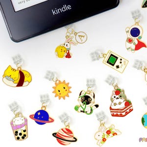 Könnte beinhalten: Eine Sammlung von 13 bunten Emaille-Anhängern mit verschiedenen Designs, darunter eine Katze, ein Raumschiff, eine Sonne, ein Planet, eine Sushi-Rolle, eine Rakete, eine Spielkonsole, ein Kaninchen, ein Boba-Tee und eine Katze auf einem Kuchen. Die Anhänger sind an durchsichtigen Plastikclips befestigt, die zum Markieren von Seiten in einem Buch verwendet werden können.
