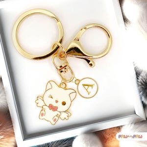 May include: Gold-coloured keychain featuring a winking white cat charm with a pink bow tie. The charm has a gold border and a small circular tag with the letter "A". The keychain includes a large ring and a lobster clasp.