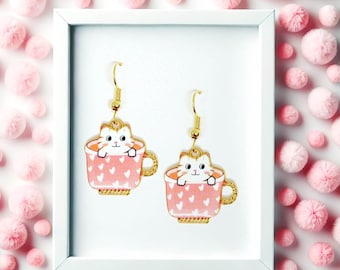 May include: A pair of gold-toned dangle earrings featuring a white cat sitting inside a pink teacup decorated with small white hearts. The earrings hang from gold-toned hooks and are displayed within a white frame against a pink background.