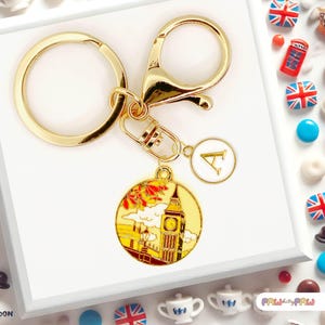 Puede incluir: Llavero dorado con un dije de Big Ben y un colgante con la inicial "A". El dije de Big Ben presenta un diseño amarillo y dorado con detalles en rojo y naranja. El llavero incluye un cierre y un anillo.