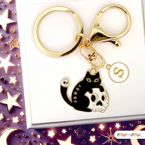 May include: Gold keyring featuring a black cat with white eyes sitting on a white skull with black eyes. The keyring also has a gold split ring and a gold charm with the letter "S" on it.