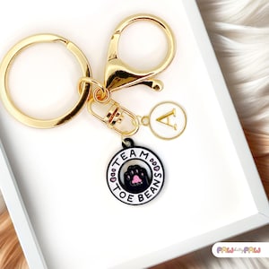 Puede incluir: Llavero dorado con un anillo y cierre grandes. Cuenta con un dije con el texto "TEAM TOE BEANS" alrededor de una huella de pata, y un dije con la letra "A". El llavero está sobre una superficie blanca.