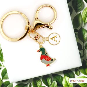 Op de afbeelding: Goudkleurige sleutelhanger met een eendenbedel. De eendenbedel is rood, groen, grijs en wit. De sleutelhanger heeft ook een ronde bedel met de letter "A". De sleutelhanger ligt op een wit oppervlak.