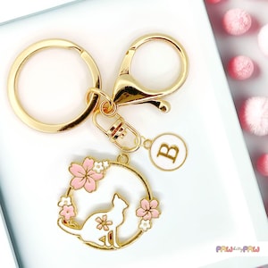 May include: Gold keyring with a white cat charm surrounded by pink and white flowers. The charm also has a gold circle around it. The keyring has a gold split ring and a gold lobster clasp. The keyring also has a small gold charm with the letter B on it.