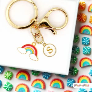 May include: Gold keyring with a rainbow charm, a small circle charm with the letter "S" and a gold lobster clasp. The keyring is displayed on a white surface with rainbow-coloured candy.