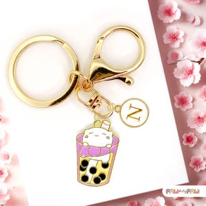 May include: Gold-tone keychain with a charm depicting a cartoon boba tea cup and a white character. The keychain also has a round charm with the letter "N" in a white circle. The background features pink flower accents.
