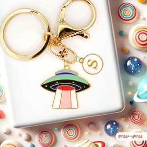Op de afbeelding: Goudkleurige sleutelhanger met een bedel met een kleurrijk UFO-ontwerp. De bedel heeft een zwart, blauwgroen en rood kleurenschema. Een extra bedel met de letter "S" is ook bevestigd. De sleutelhanger wordt gepresenteerd op een wit oppervlak met snoep.