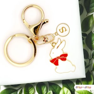 May include: Gold keyring with a white rabbit charm with a red bow and a gold circle charm with the letter "S" on it.