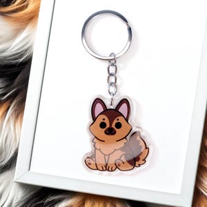 Puede incluir: Un llavero acrílico transparente con un diseño de perro pastor alemán de dibujos animados. El perro está representado en tonos marrones, beige y negros, con una cola y orejas esponjosas. El llavero tiene un anillo y una cadena de metal plateado.