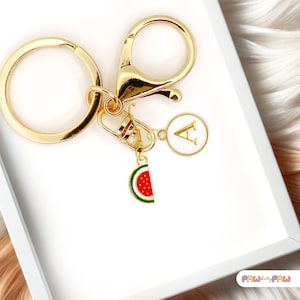May include: Gold-toned keychain with a lobster clasp, key ring, and charms. Charms include a white circle with the letter "A" and a watermelon slice with red seeds and green rind. The keychain is on a white surface.