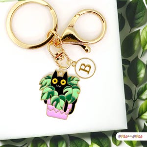 Könnte beinhalten: Goldener Schlüsselanhänger mit einer schwarzen Katze, die in einem rosafarbenen Topf mit grünen Blättern sitzt. Der Schlüsselanhänger hat auch einen goldenen Anhänger mit dem Buchstaben B.