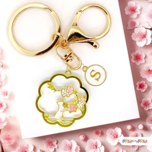 May include: Gold keychain with a charm featuring a white cat sitting in a pink and white cherry blossom tree. The charm is attached to a gold split ring and a gold lobster clasp. The keychain also has a small gold charm with the letter "S" on it.