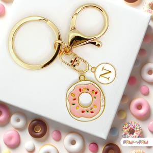 May include: Gold keyring with a pink and white donut charm and a gold charm with the letter N. The keyring is surrounded by colourful sprinkles.