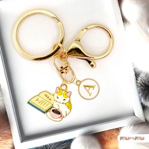 May include: Gold-coloured keychain featuring a cat charm reading a book and drinking coffee. The charm is white, yellow, and pink. A round charm with the letter "A" is also attached. The keychain has a clasp and a large ring.
