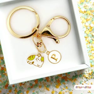 May include: Gold-coloured keychain with a lobster clasp and a round key ring. The keychain features two charms: a white and green cat charm and a white circular charm with the letter "P" in gold. The keychain is presented in a white box.