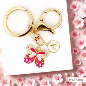 Puede incluir: Llavero dorado con un dije de mariposa y un colgante circular con la letra "A". El dije de mariposa presenta un diseño blanco y rosa. El llavero está sobre un fondo de flores rosas.