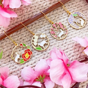 Peut inclure: Quatre boucles d'oreilles dorées avec un lapin blanc et différents motifs floraux. Les motifs comprennent une fleur de camélia, des fleurs de cerisier, du glycines et des fleurs de prunier. Les boucles d'oreilles sont sur une surface en bois avec des fleurs roses.