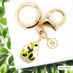 May include: Gold keychain with a charm of a terrarium with green and black succulents. The keychain also has a small gold charm with the letter "S" on it.
