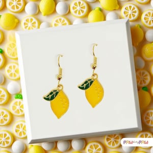 Puede incluir: Un par de pendientes de gancho en tono dorado con dijes de limón amarillo y hojas verdes. Cada dije de limón está elaborado con detalles en esmalte, mostrando un cuerpo de fruta amarillo brillante y una hoja verde en la parte superior.