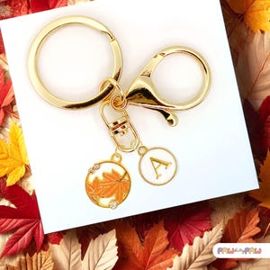 May include: Gold-toned keychain with a lobster clasp and a round key ring. The charm features a white enamel circle with the letter "A" and a round charm with orange maple leaves and clear crystals. The background has autumn leaves.