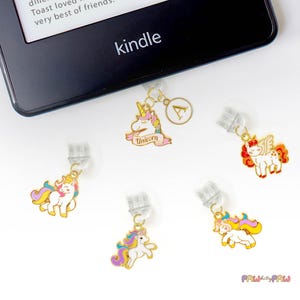 Könnte beinhalten: Ein schwarzer Kindle-E-Reader mit mehreren bunten Charms zum Thema Einhorn. Die Charms haben goldfarbenes Metall mit Emaille-Details in Rosa, Gelb, Lila und Weiß. Ein Charm hat den Buchstaben "A". Das Wort "Unicorn" steht auf einem rosa Banner.