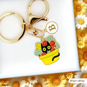 May include: Gold keyring with a black cat peeking out of a yellow pot with red and green flowers. The keyring also has a gold letter B charm.