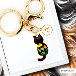 May include: A gold-coloured keyring with a black cat-shaped charm. The cat charm has a floral design with yellow, pink, and green accents. The keyring also includes a gold ring, clasp, and a charm with the letter "A".