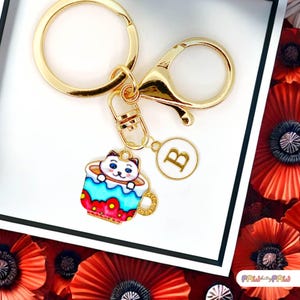 Llavero esmaltado con diseño de gato blanco en taza de té con flores azules y monograma personalizado. Divertido llavero kawaii bañado en oro - K-A006-7