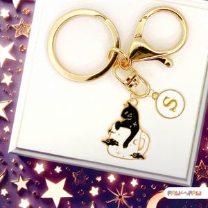 May include: Gold keyring with a black cat sitting in a white mug with stars and clouds. The keyring also has a gold circle charm with the letter "S" on it.