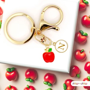 Op de afbeelding: Gouden sleutelhanger met een rode appelbedel en een gouden cirkelbedel met de letter N. De sleutelhanger is omringd door kleine rode appelbedels.