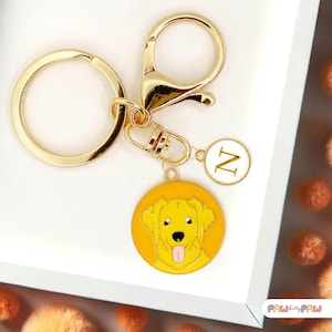 Puede incluir: Un llavero dorado con un dije redondo que presenta un perro de dibujos animados amarillo con la lengua fuera. El dije está rodeado por un borde amarillo. Un dije blanco más pequeño con la letra "N" cuelga cerca.