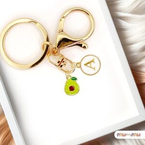 Peut inclure: Porte-clés doré avec un mousqueton, un anneau rond et un charm avec la lettre "A". Le porte-clés comprend également un charm avocat vert avec une graine rouge. Le fond est blanc.