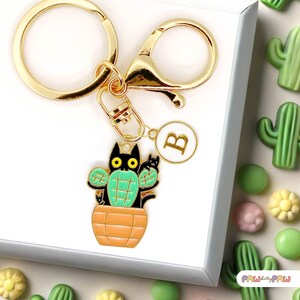 Puede incluir: Llavero dorado con un dije de gato negro cactus. El dije presenta un gato negro con ojos amarillos y brazos de cactus verdes, sentado en una maceta naranja. Incluye un dije redondo con la letra "B".