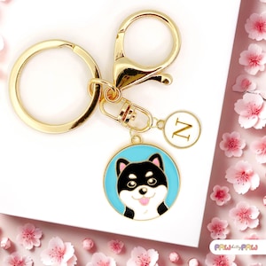 Puede incluir: Llavero dorado con un charm circular azul que presenta un perro Shiba Inu negro y blanco con la lengua rosa. El charm también tiene un charm de oro con la letra N.