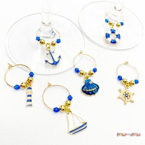 Può includere: Set di sei ciondoli per bicchieri da vino a tema nautico. Ogni ciondolo presenta un design diverso, tra cui un'ancora, un faro, una barca a vela, una conchiglia, un timone e un salvagente. I ciondoli sono in metallo dorato con dettagli in smalto blu e bianco.