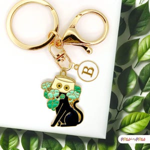 May include: Gold keyring with a black cat charm wearing a white hat with green flowers. The cat has yellow eyes and the keyring has a gold letter 'B' charm.