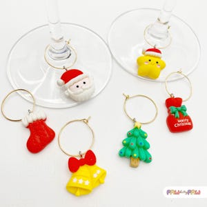 May include: A collection of Christmas-themed wine charms. The charms include a red stocking, a yellow bell with stars, a green Christmas tree, a red gift bag, a yellow star, and a Santa Claus head. Each charm hangs from a gold-colored ring.