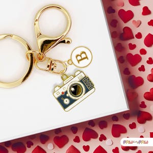 Peut inclure: Porte-clés en or avec un charme en forme d'appareil photo, un charme en forme de lettre B et un anneau brisé en or. Les breloques sont sur un fond blanc avec des cœurs rouges.