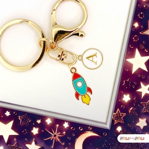 May include: Gold-toned keychain with a rocket charm and a circular charm with the letter "A". The rocket is red, blue, and yellow. The keychain is on a white surface with a decorative border featuring stars and moons.