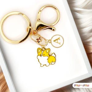 Puede incluir: Llavero dorado con un dije de corgi y un dije circular con la letra "A". El dije de corgi es amarillo y blanco con contornos negros. El llavero tiene un anillo grande y un mosquetón.
