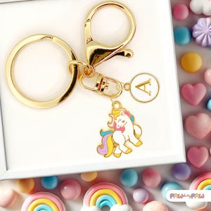 Peut inclure: Porte-clés doré avec un charm rond avec la lettre "A" et un pendentif licorne coloré. La licorne a un corps blanc, un nœud rose et une crinière et une queue arc-en-ciel. Le porte-clés est entouré de bonbons colorés.