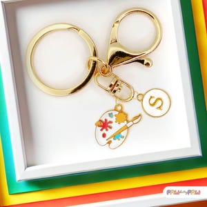 May include: Gold keychain with a paint palette charm, a paintbrush, and a letter 'S' charm. The keychain is attached to a gold key ring.