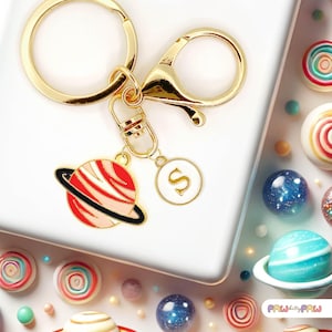 May include: A gold-coloured keyring with a planet charm featuring red and white stripes, a black ring, and a white circle charm with the letter "S". The keyring is attached to a gold clasp. The background includes colourful sweets and decorative spheres.