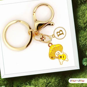May include: Gold-coloured keyring with a round clasp and a charm featuring a yellow mushroom playing a yellow guitar. The keyring also includes a white charm with the letter "B".